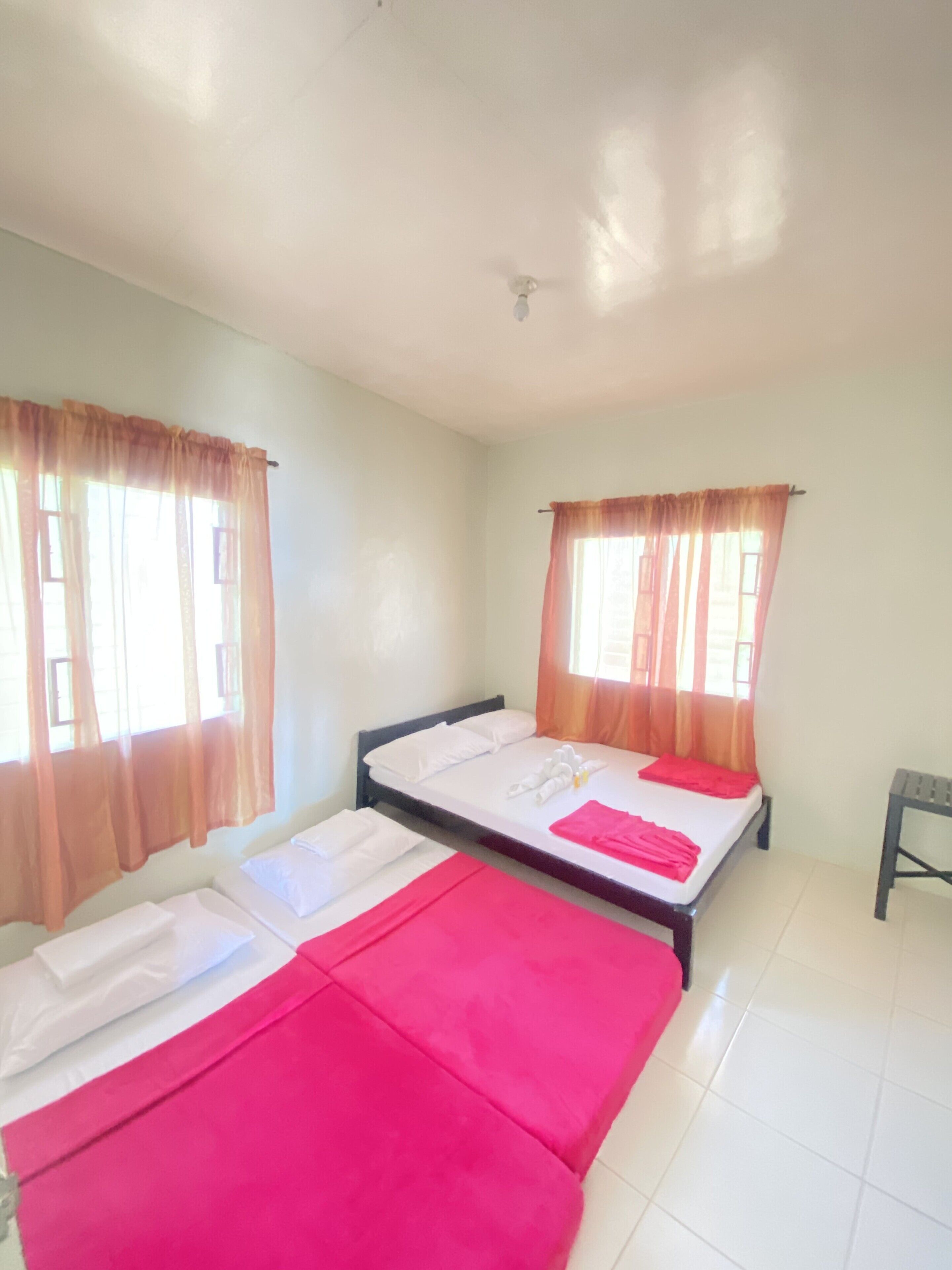 A room with two beds, each with white linens and a red blanket. There are windows with orange curtains, and a small table is visible in the corner.
