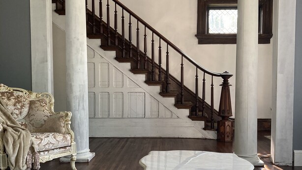 A spacious living room with a grand staircase, a plush sofa, and a marble coffee table. - 1888 Victorian House (Mitchell)