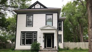 A two-story white house with a black front door, a small porch, and a number '215' on the front wall. - 1888 Victorian House (Mitchell)