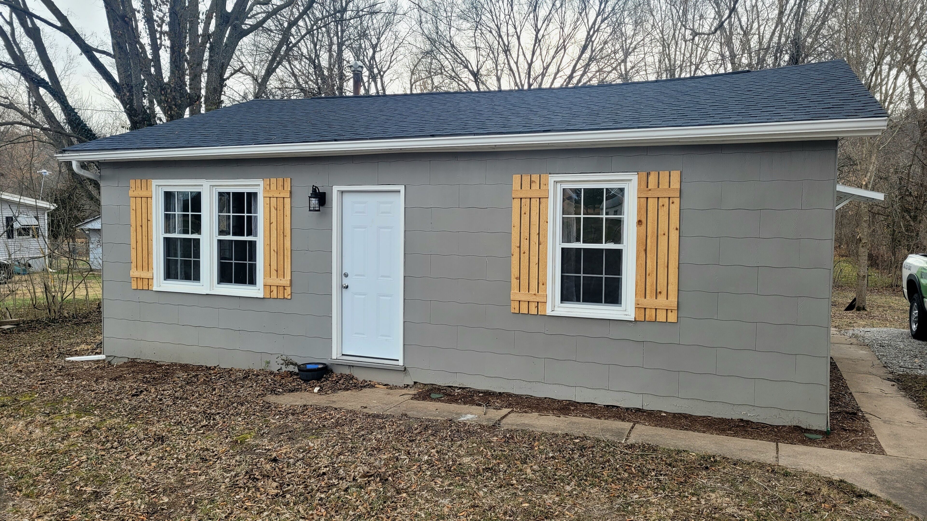 A small, single-story house with a gray exterior, two windows with wooden shutters, and a white door.