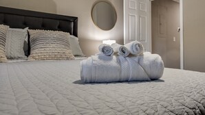 Un lit bien fait, recouvert d’une courtepointe matelassée, une pile de serviettes enroulées et un miroir rond accroché au mur.