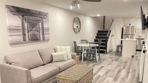 Una sala moderna con un sofá gris, un reposapiés de mimbre, una mesa de comedor con sillas, un ventilador de techo y una escalera que conduce a una zona de cocina.