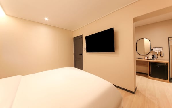 A hotel room with a bed, a TV mounted on the wall, a mirror, and a small desk with a hairdryer and other items.
