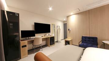 A modern hotel room with a bed, a desk with a computer, a chair, a wardrobe, and a mirror.
