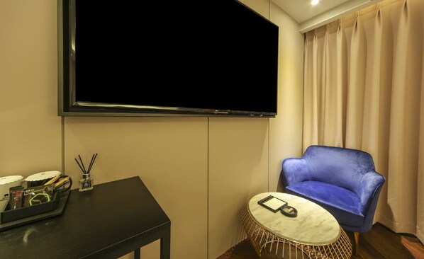 A hotel room with a large flat-screen TV mounted on the wall, a blue armchair, a small round table with a white marble top, and a tray with various items on a black table.