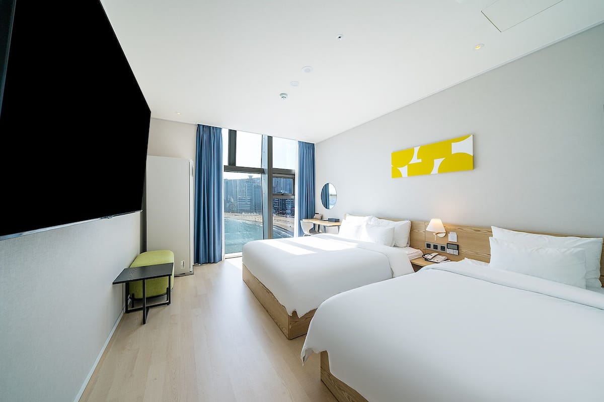 A modern hotel room with two beds, a desk, and a view of the city.