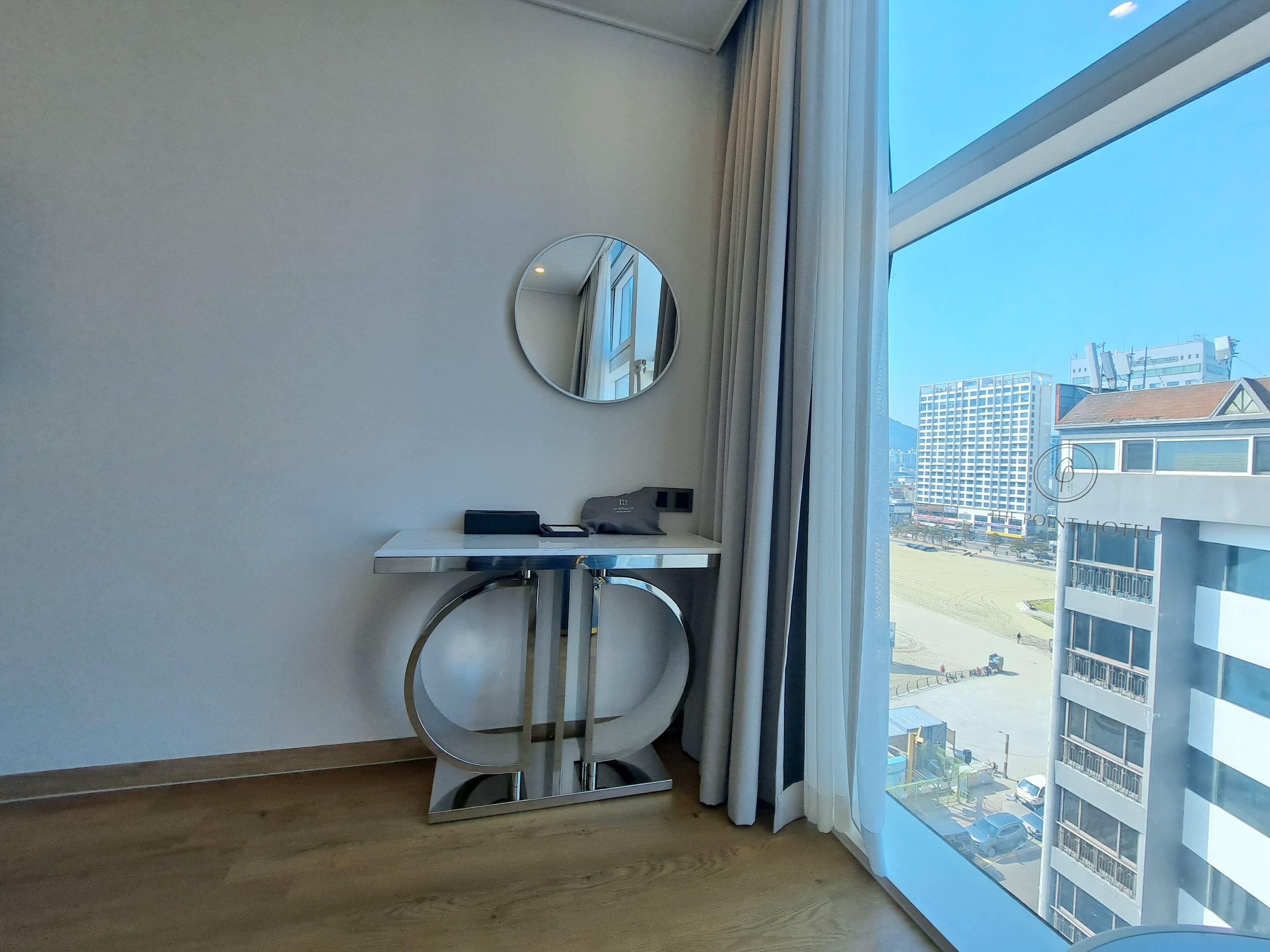 Habitación de hotel con un ventanal grande, un espejo redondo, una mesita pequeña y vistas de la ciudad.