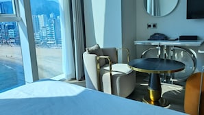 A hotel room with a bed, a round table, a chair, a small desk, a mirror, and a view of a cityscape through large windows. - Ocean the point Hotel (Busan)