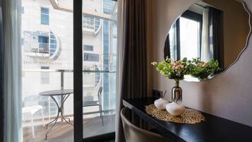 A hotel room with a large window, a round mirror, a vase with flowers, and a small table with a chair.