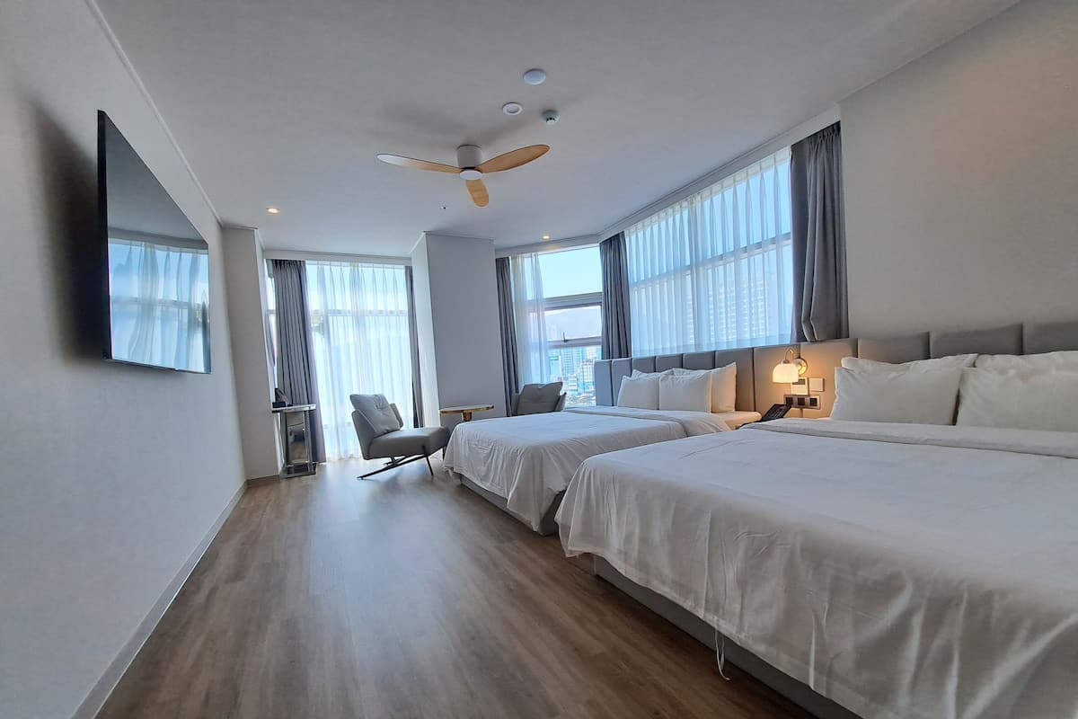 A hotel room with two beds, a ceiling fan, a TV mounted on the wall, and large windows with curtains.