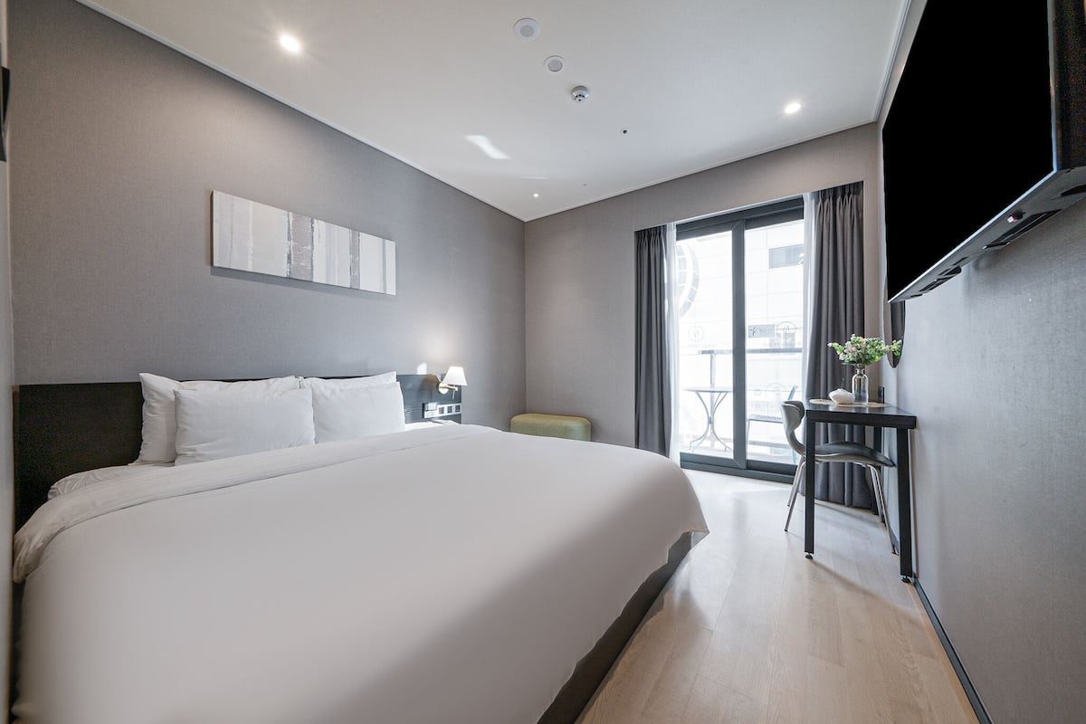 A modern hotel room with a large bed, a desk with a chair, a TV, and a window with curtains.