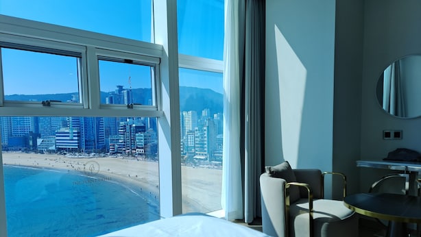 A hotel room with a large window overlooking a beach and cityscape. - Ocean the point Hotel (Busan)