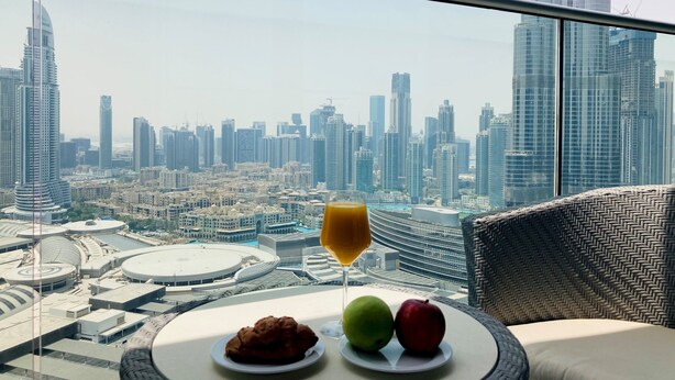 A balcony with a table set for breakfast, featuring a glass of orange juice, two apples, and a plate of pastries. The view includes a cityscape with modern skyscrapers. - Ultimate Luxury Address Boulevard Dubai - 3 Bedrooms (Dubai)