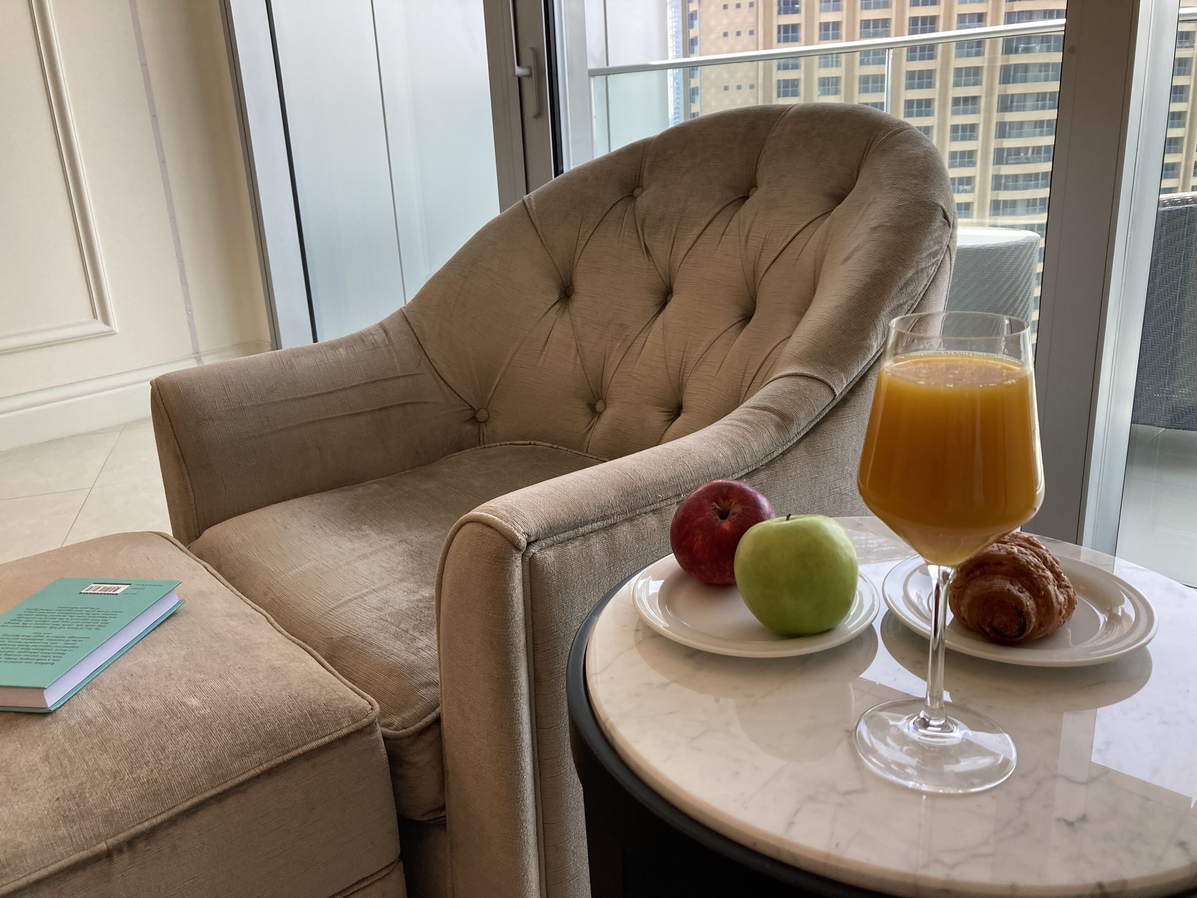 A beige armchair, a small table with a glass of orange juice, two apples, and a croissant.
