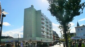 A modern building with multiple floors and balconies, surrounded by a pedestrian area with shops and a tree. - Sievers 37, Deutschland (Sylt)