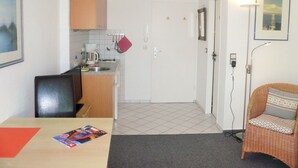 A compact room with a kitchenette, a dining table with a magazine, a wicker chair, and a framed picture on the wall. - Sievers 37, Deutschland (Sylt)