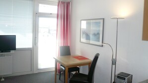 A room with a dining table, chairs, a television, a radiator, a window with blinds, a lamp, and a small kitchen area. - Sievers 37, Deutschland (Sylt)