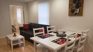 A dining area with a white table and chairs, a black sofa with red cushions, a small white side table, and a painting on the wall.