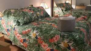 A bedroom with a bed covered in tropical floral bedding, two bedside lamps, a large mirror, and a painting on the wall.