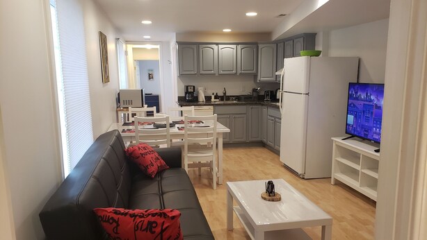 A modern living room with a black sofa and red cushions, a white coffee table, and a TV on a stand. The kitchen area is visible with grey cabinets, a white refrigerator, and a dining table with chairs.
