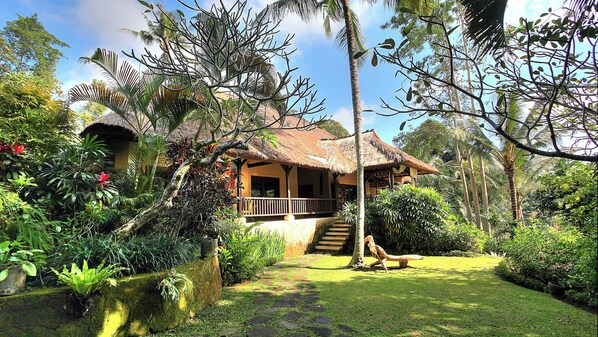 A thatched-roof house with a porch, surrounded by lush tropical vegetation and a clear sky.