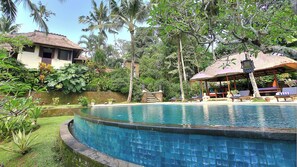 A pool surrounded by lush greenery, a thatched-roof structure, and a building with a sloped roof.
