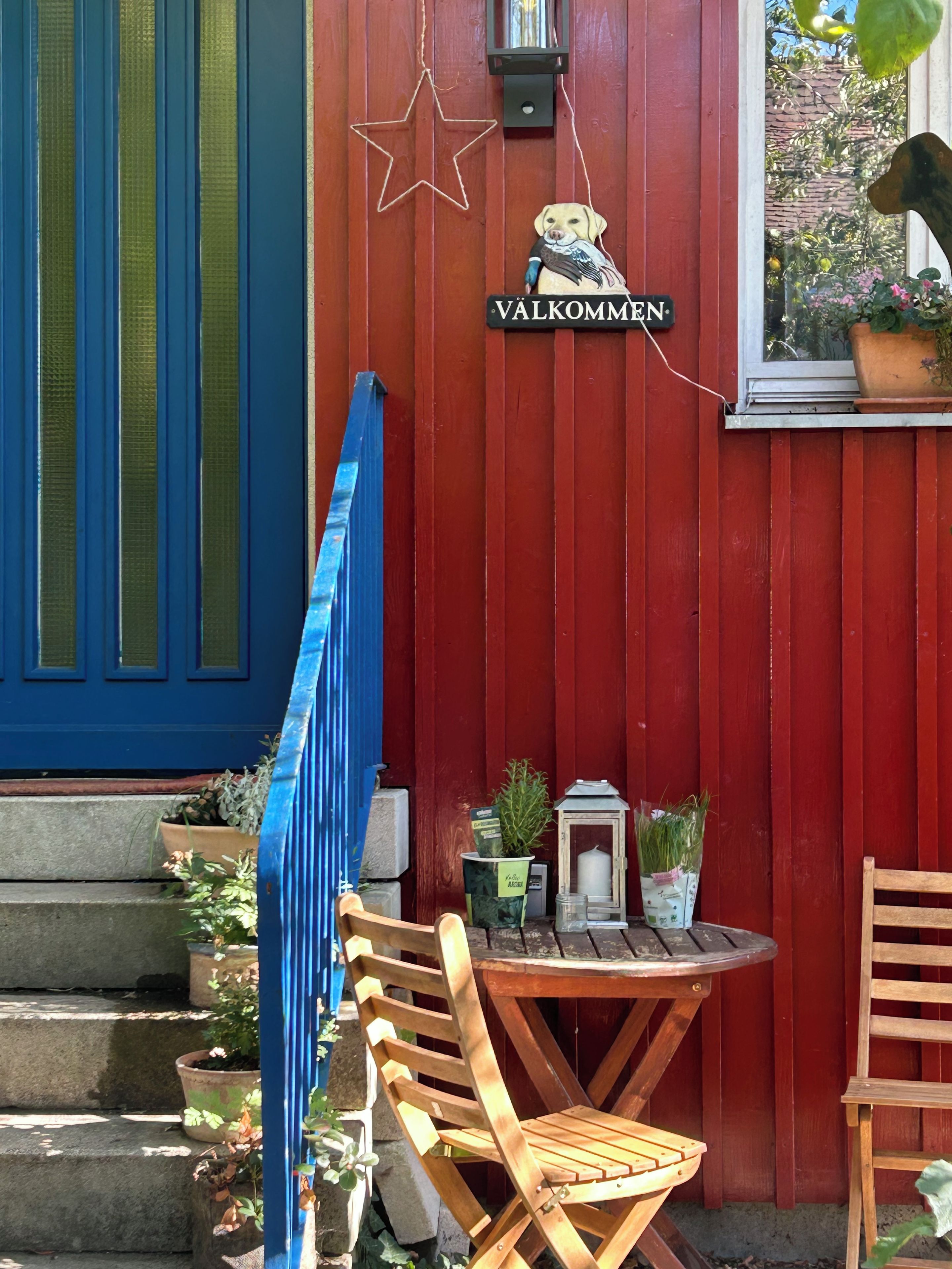 Una pared de madera roja con un letrero que dice 'VÄLKOMMEN' y una decoración de estrella. Hay una pequeña mesa con sillas, un farol y plantas en macetas.