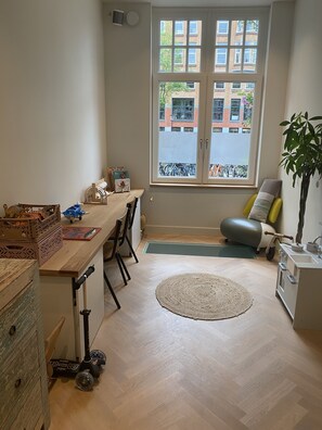 A room with a wooden desk, chairs, a small rug, a window with a view of buildings, and a plant.
