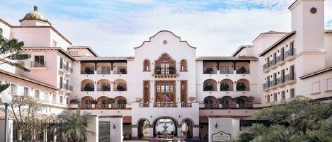 A large, white building with a central entrance, multiple balconies, and a courtyard with plants and seating.