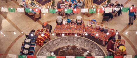 Un área de comedor al aire libre con mesas, sillas y sombrillas. Se observa un escenario con un artista y una piscina al fondo.