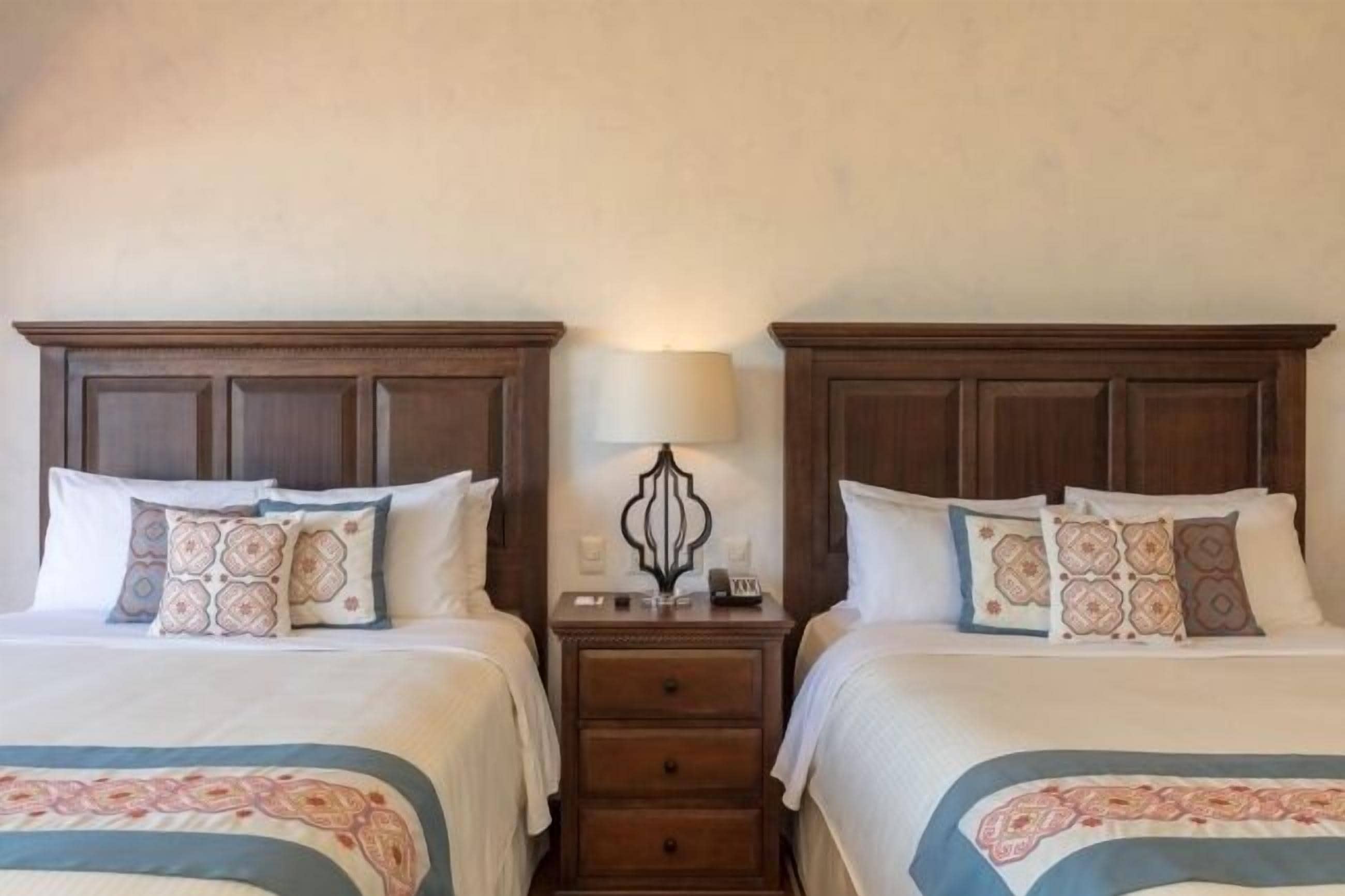 A hotel room with two beds, each with a wooden headboard, white bedding, and patterned pillows. There is a nightstand with a lamp and a phone between the beds.