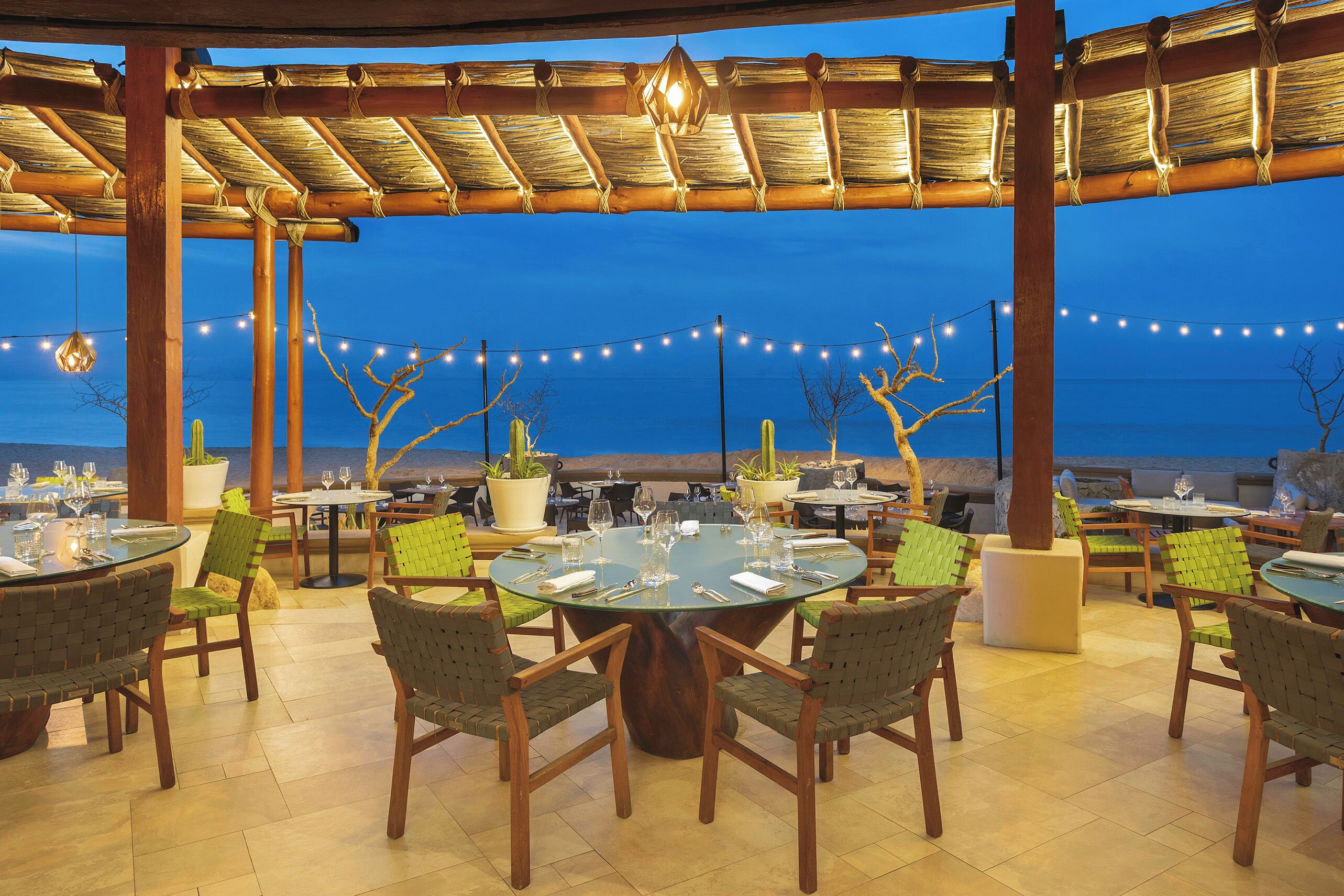 Un espacio de comedor al aire libre con mesas y sillas redondas, luces colgantes y plantas en macetas.