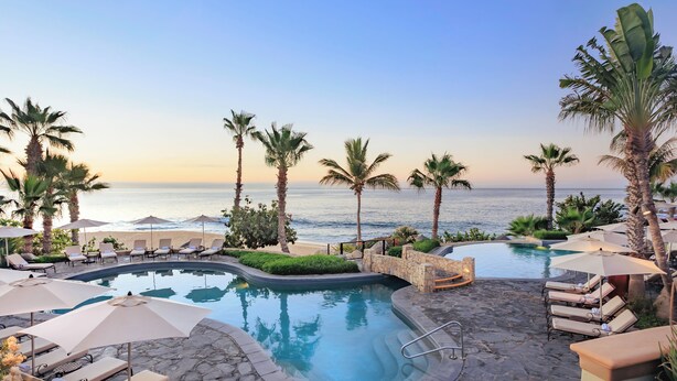 A pool area with lounge chairs and umbrellas, surrounded by palm trees and a view of the ocean.