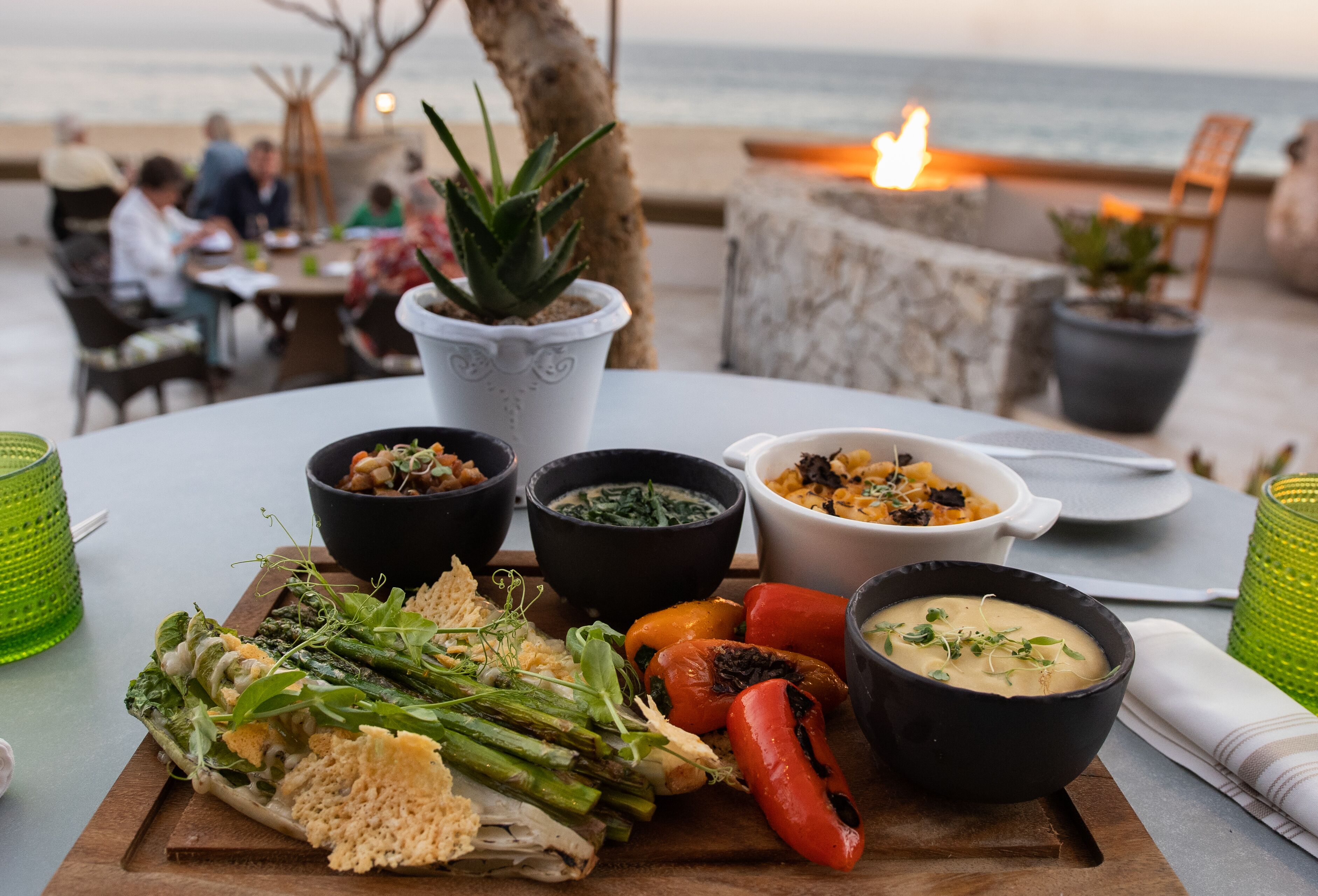 Ein Tisch mit einer Auswahl an Speisen, darunter Gemüse, Brot und Suppenschalen, draußen mit Blick auf das Meer, im Hintergrund essen Menschen.