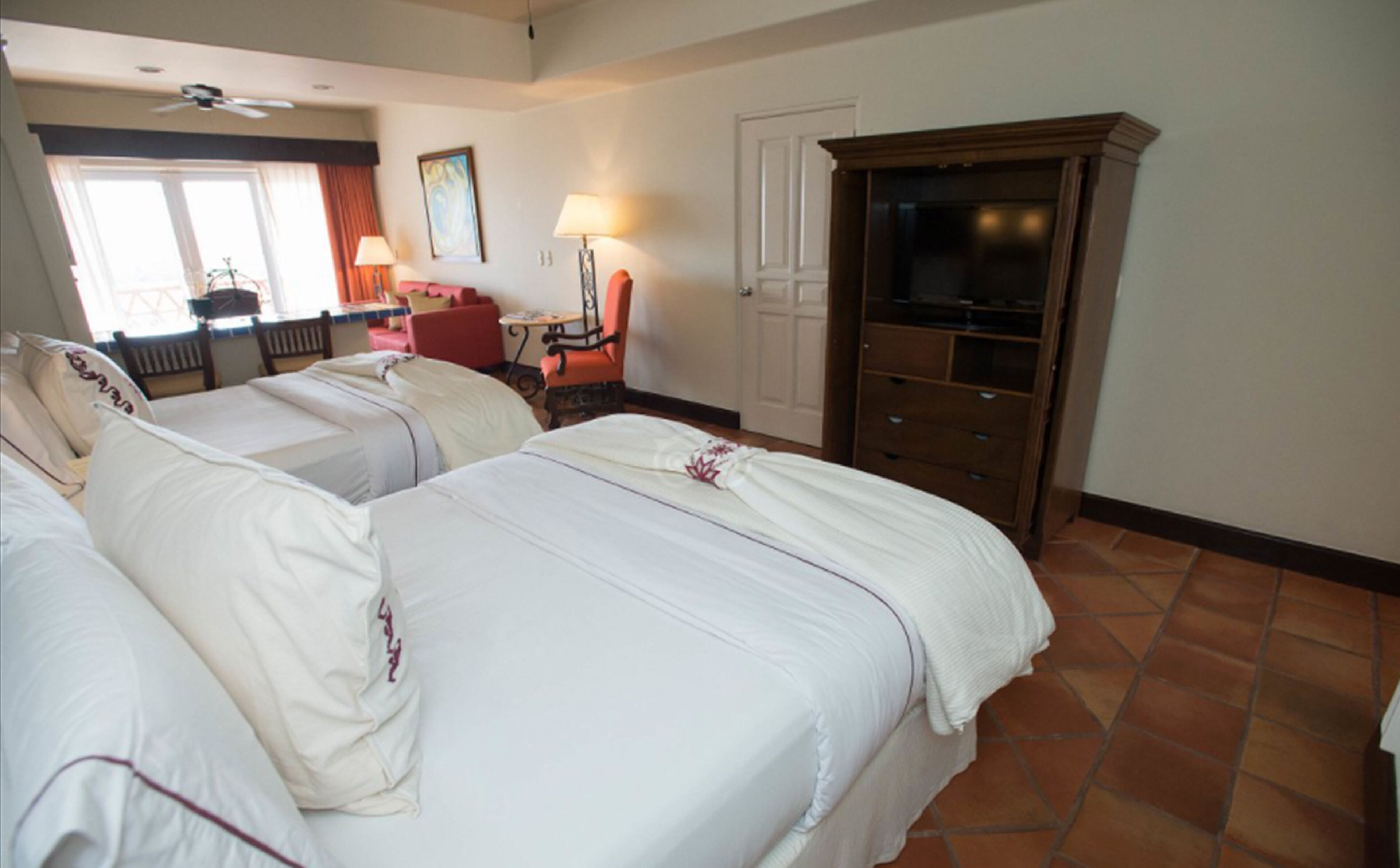 A hotel room with two beds, a television, a desk with a lamp, and a chair.