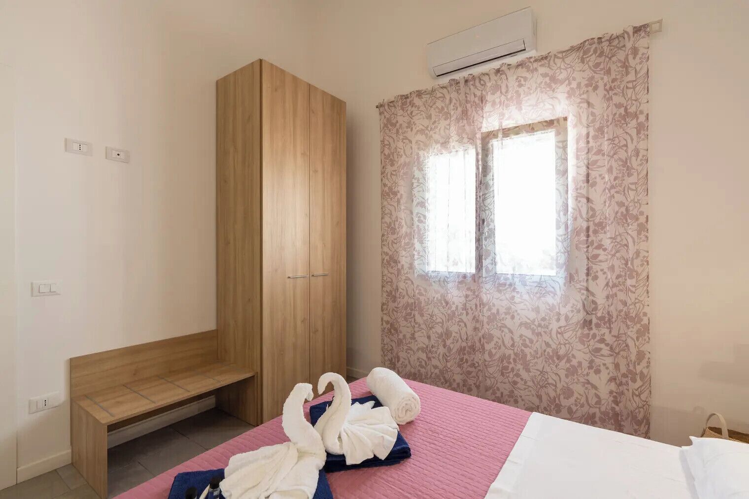 A bedroom with a wooden wardrobe, a bench, a bed with pink and white bedding, and a window with floral curtains.