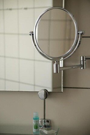 Une salle de bain avec un grand miroir rond fixé au mur, une petite étagère contenant des articles de toilette et une fenêtre avec des stores.