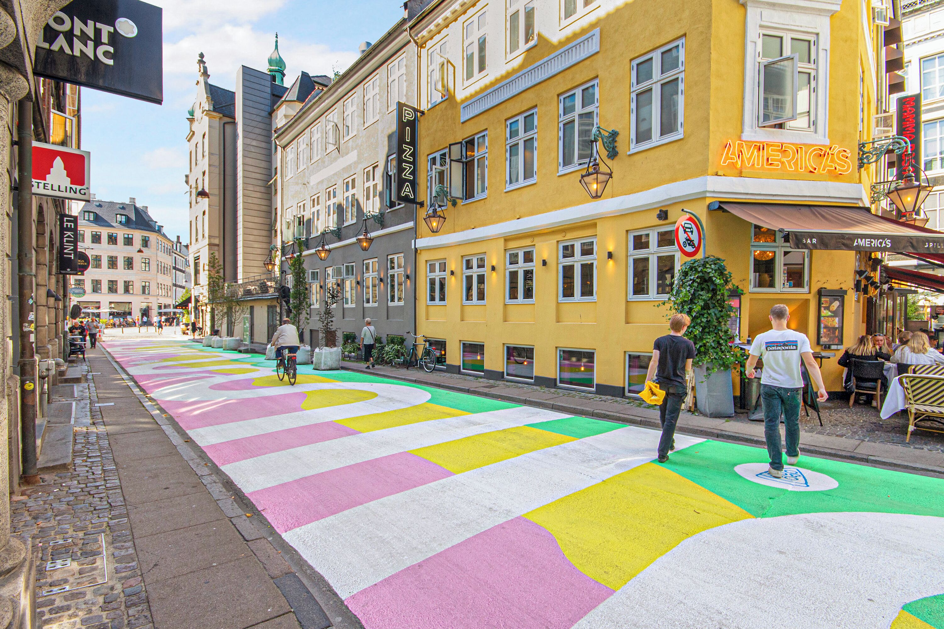 Una calle con carriles bici coloridos, un edificio amarillo con un cartel que dice 'AMERICAS' y un cartel de 'PIZZA' en un edificio.