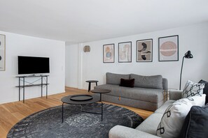 Een moderne woonkamer met een grijze bank, een zwarte salontafel en een flatscreen televisie op een standaard.