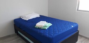 Una cama con colchón y almohadas azules, una manta doblada y una ventana pequeña al fondo.