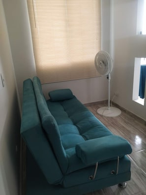 A teal sofa in a room with a standing fan, wooden flooring, and a window with blinds. - Total Spacio Relax in Cundinamarca (Tocaima)