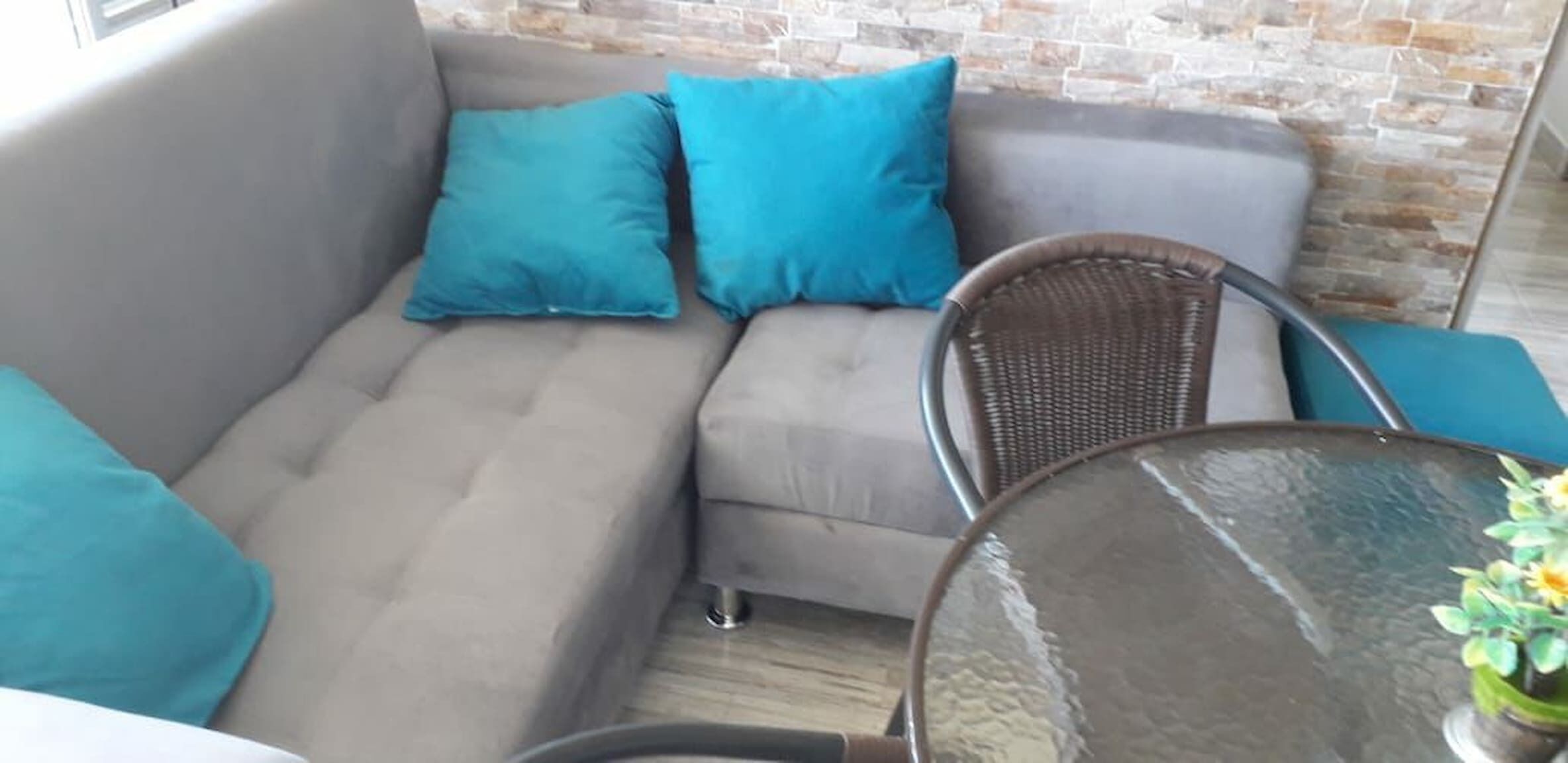 Un sofá gris con dos cojines verde azulado, una silla de mimbre y una pequeña mesa redonda con un jarrón de flores.