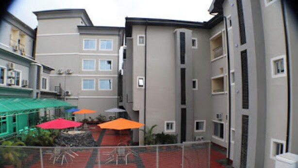 A multi-story residential building with balconies, a covered outdoor seating area with umbrellas, and a red-tiled courtyard. - Room-all Seasons Hotel - Business Suite (Owerri)