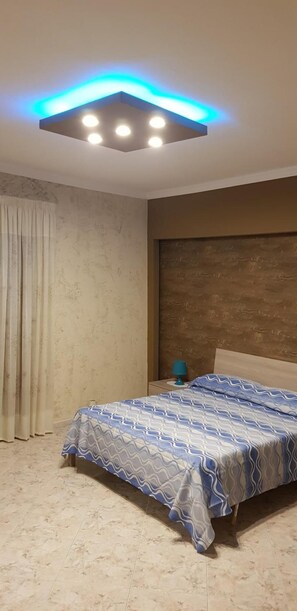 Un dormitorio con cama, mesita de noche, cabecero fijado a la pared y lámpara en el techo.