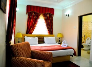 A hotel room with a bed, two bedside lamps, a chair, a window with curtains, and a bathroom visible through an open door. - Room-auris Court - Guest Room (Abuja)