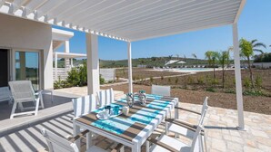 A covered outdoor dining area with a table set for two, a white chair, and a view of a pool and landscape.