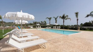 A pool area with white lounge chairs and umbrellas, palm trees, and a clear sky.