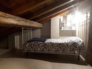 A small, dimly lit attic bedroom with a single bed, wooden beams, and a window with a view of the outside.