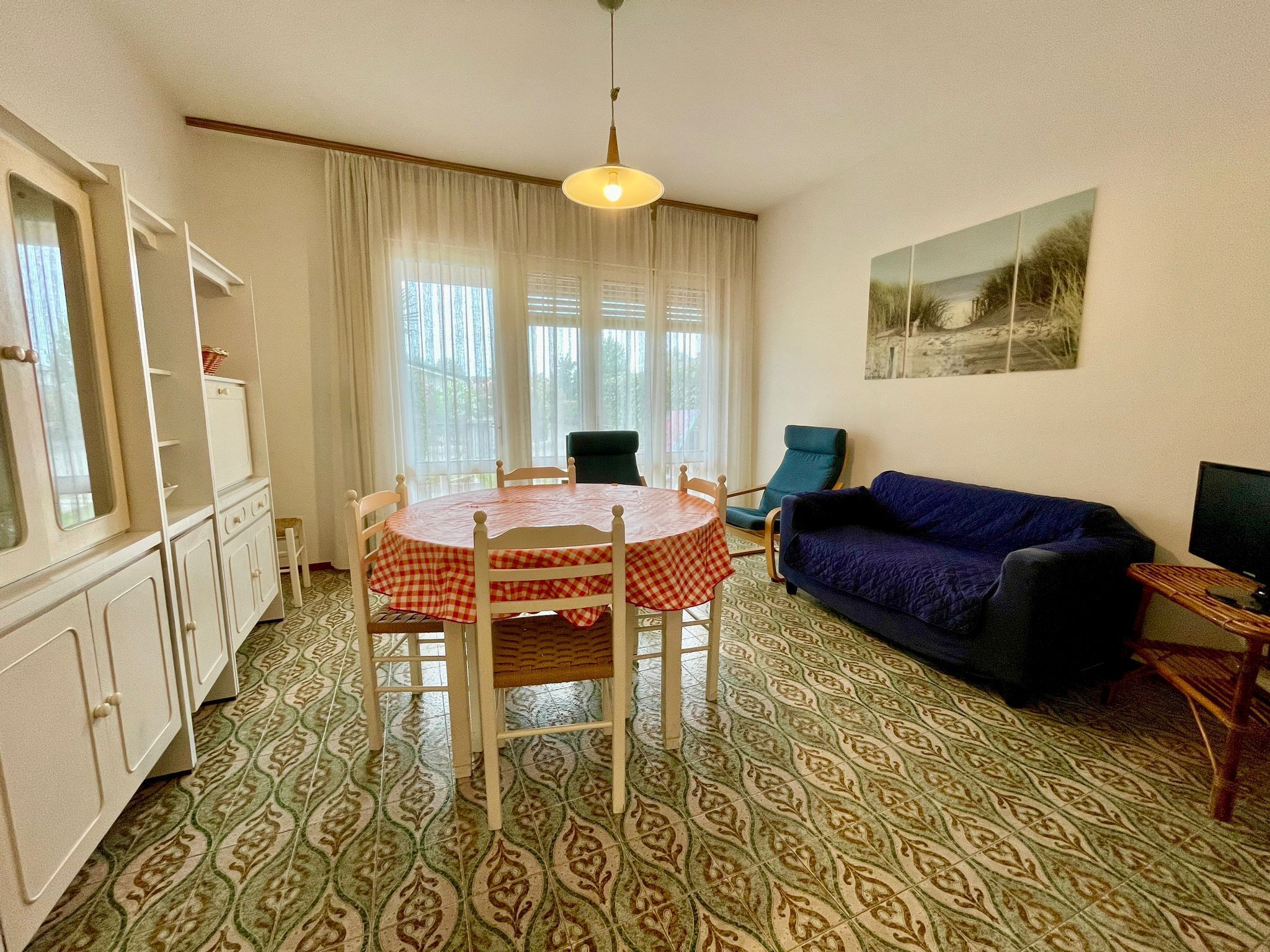 A room with a round table covered by a checkered tablecloth, a blue sofa, a television on a wooden stand, and patterned floor tiles.