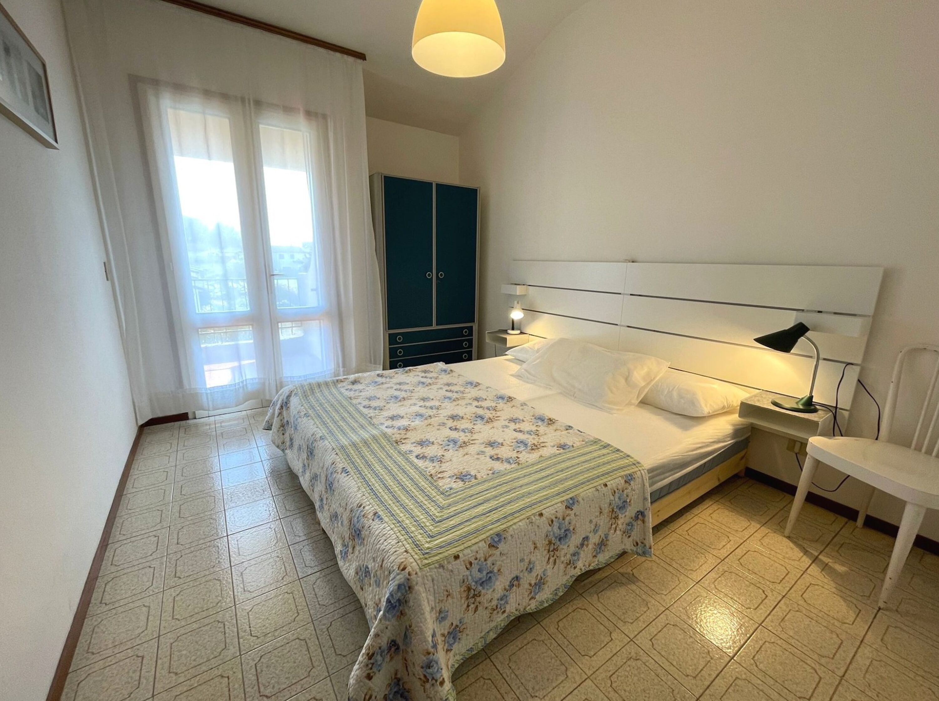 A bedroom with a bed, a nightstand, a lamp, a chair, a wardrobe, and a window with curtains.